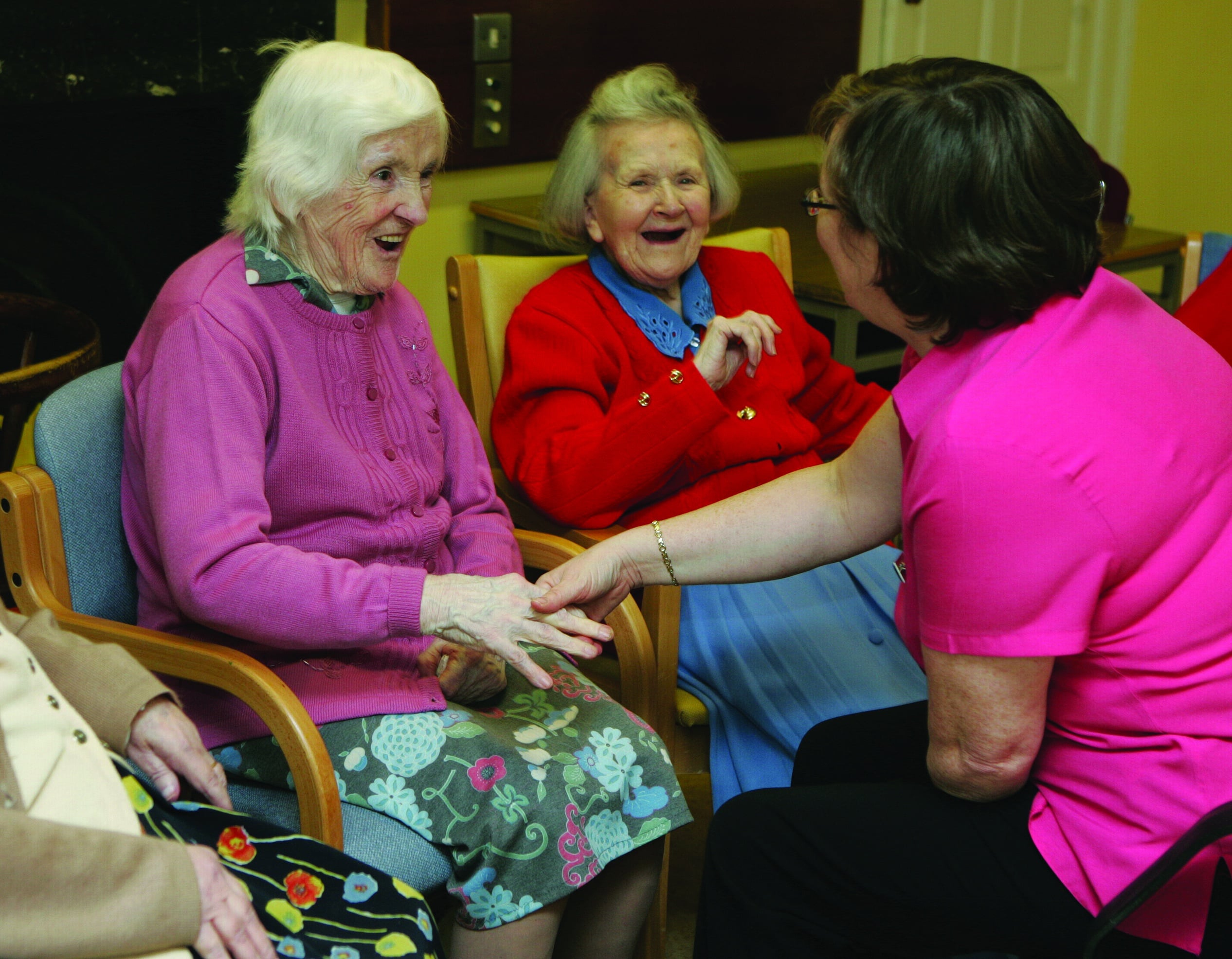 A woman in a pink shirt engages in conversation with two older women, sharing smiles and attentive expressions.