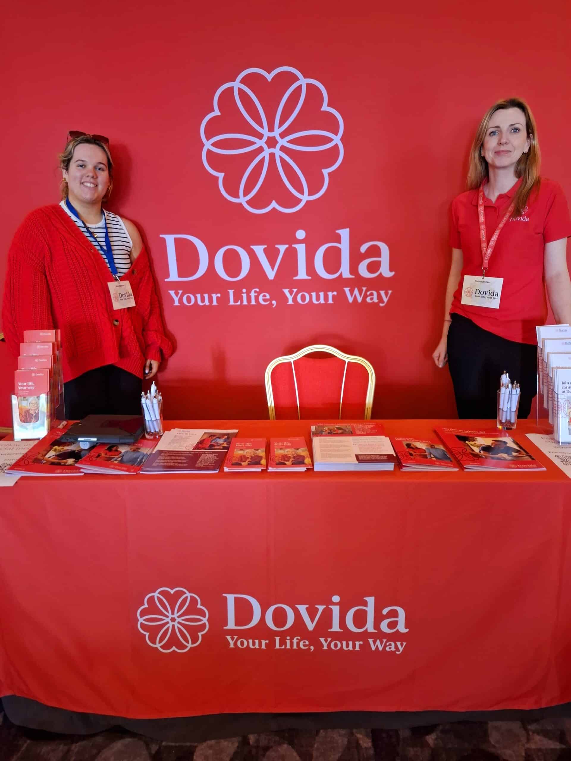 Two women stand behind a table displaying a sign that reads "Dovida," smiling and engaging with attendees.
