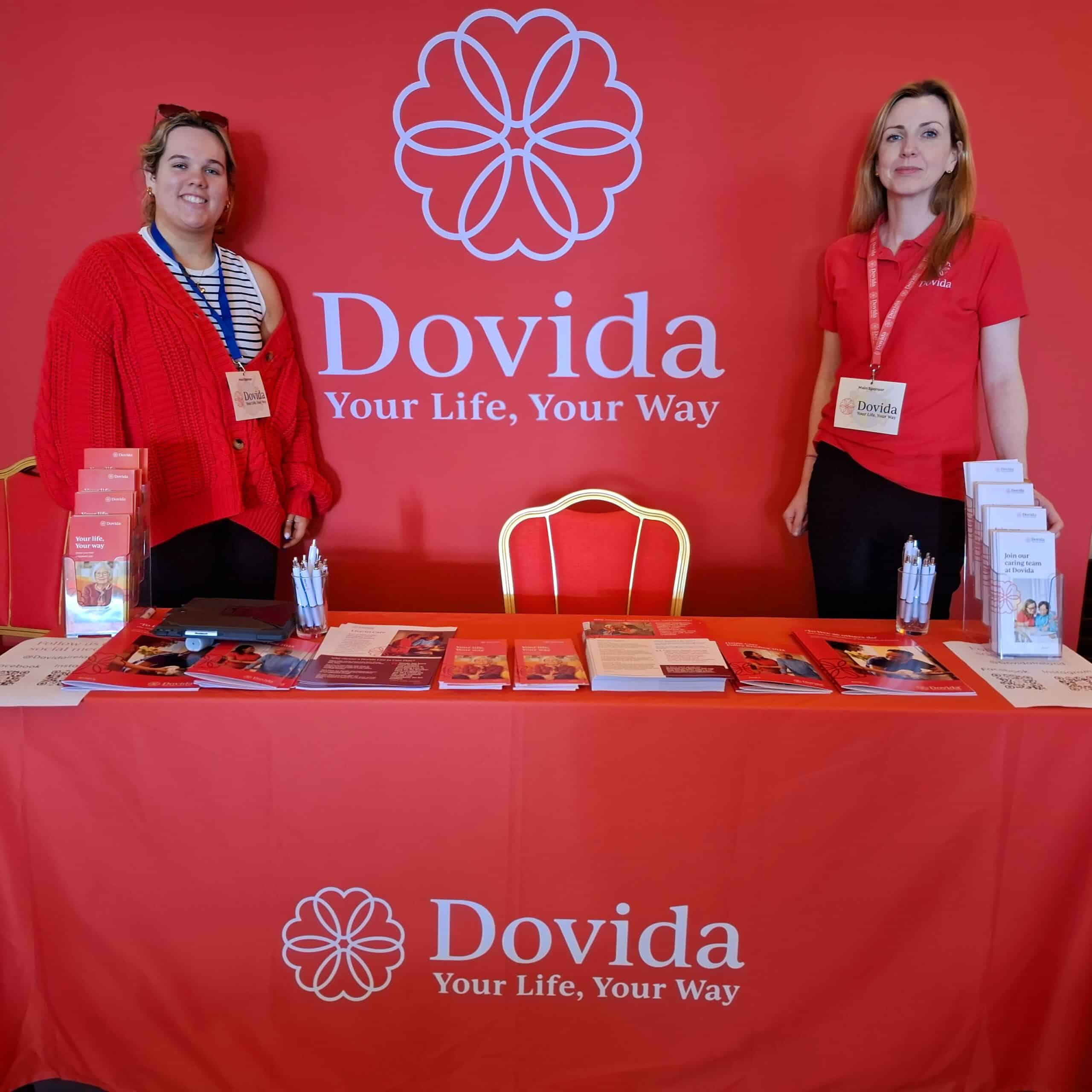 Two women stand behind a table displaying the word "Dovida" prominently.